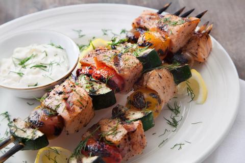 Salmon and vegetable kebabs covered in herbs next to a small dish of white sauce