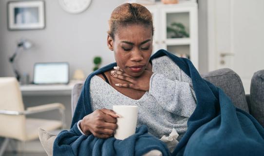 Woman wrapped in blanket on couch, holding hot beverage, hand on sore throat