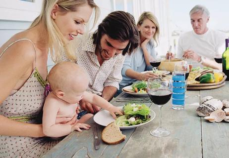 Two parents sit with their baby at the dinner table that has salad and wine glasses on it