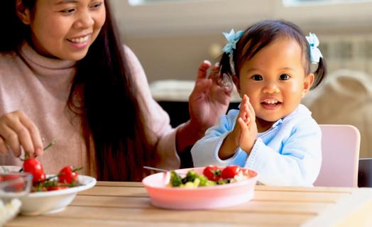 Caregiver with happy toddler at the table eating veggies