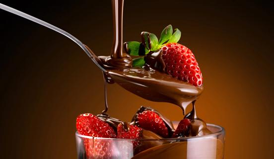 Spoon with a strawberry, with chocolate drizzling down over it, and drizzling down over more strawberries in a glass dish