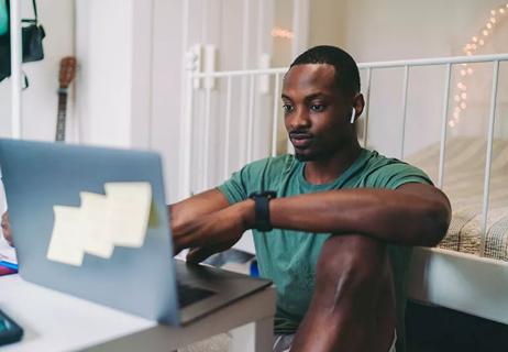 A man wearing a green shirt and sitting at his laptop working while wearing white earbuds