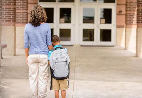 Pictured from behind, parent and nervous child stand outside of school doors.