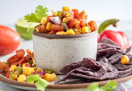 Small bowl of corn salad with red peppers and shallots on a plate, surrounded by tortilla chips