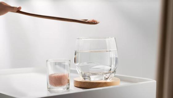 Wooden spoon with pink Himalayan salt over glass of water, with container of pink Himalayan salt