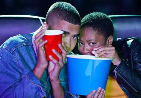 A couple huddling in their seats at a scary movie, hiding behind their drink and popcorn