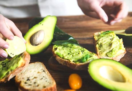 Person making avocado toast, showing avocado with seed in background.