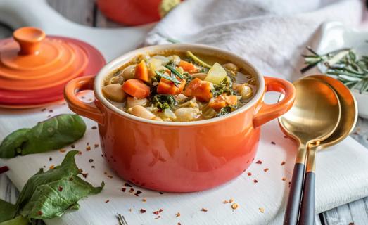 A bright orange ceramic pot full of white bean stew with rosemary and spinach, with spoons and crock lid on table