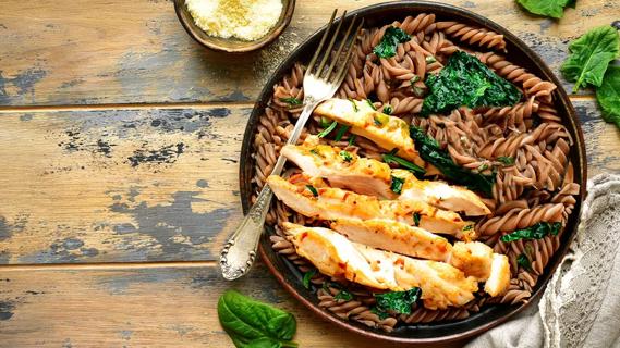 Plate  of grilled chicken on a bed of wheat pasta and cooked spinach on a wooden table