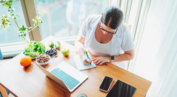 Person journaling at desk in sunny home office, with laptop, smartphone and healthy fruits and nuts on desk