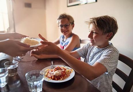 A kid with a plate of spaghetti in front of them being handed a bowl of shredded cheese