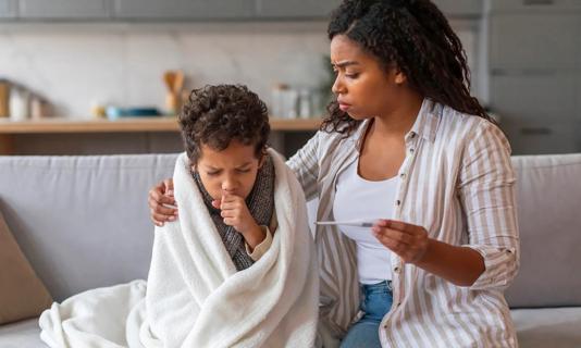 Caregiver holding a thermometer, comforting sick child wrapped in blanket on couch