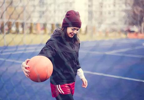 Young woman wears a windbreaker and beanie while playing basketball on an outdoor court