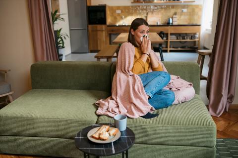 Person sitting on couch at home, wrapped in blanket, blowing their nose, with toast and tea on table