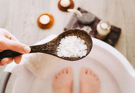 A person stands barefoot in a small tub full of water and holds a spoon of epsom salt.