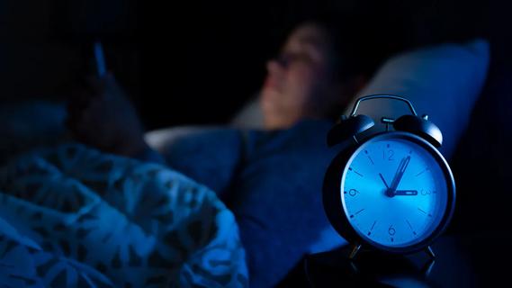 A person reading in bed in the dark with an alarm clock in the foreground.