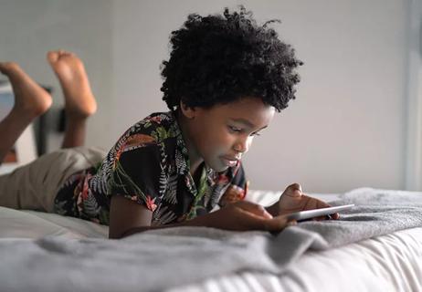 A child laying on their stomach in bed holding a tablet computer