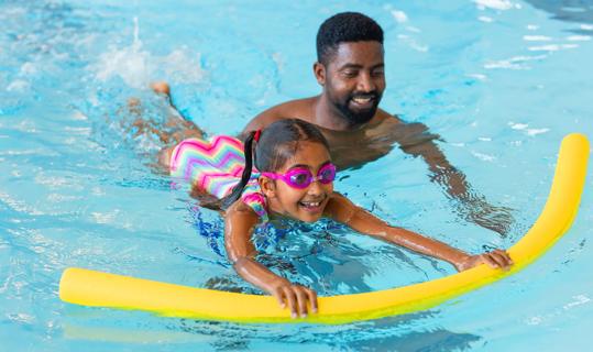 A child uses a pool noodle to stay afloat while an adult supervises behind them