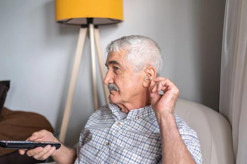 Older person holding TV remote, tugging on ear, trying to hear