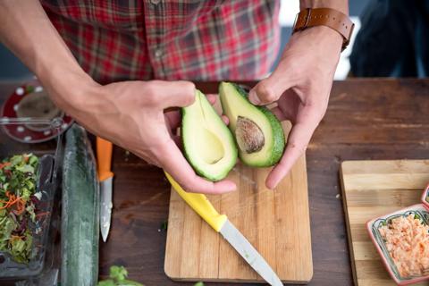 Person holding sliced open avocado halves over cutting board, among other food prep