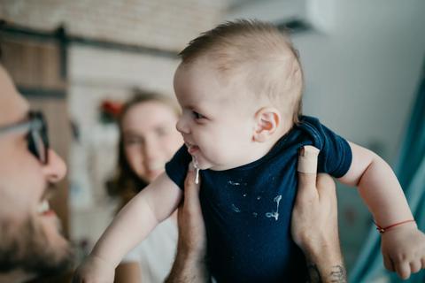 Smiling caregiver holding up smiling baby with spit-up on face and onsie, with smiling caregiver in background, too