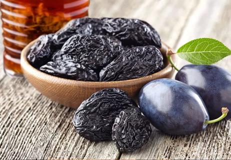A wooden bowl of prunes with prune juice in background and fresh plum in the foreground.