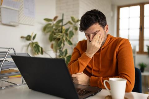 Tired person sitting at their laptop with hand rubbing their eyes, with cup of coffee nearby
