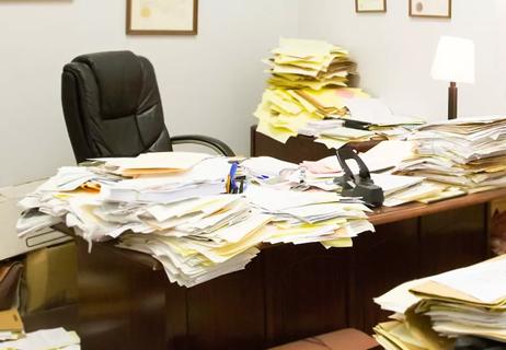 Workplace office desk filled with stacks of cluttered paperwork.