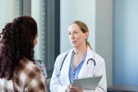 Healthcare provider, with stethoscope around neck, holding clipboard, talking to person in their office