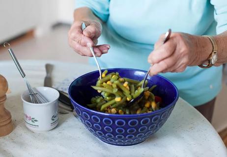 Elderly woman's hands toss bean salad in bowl on a white kitchen table.