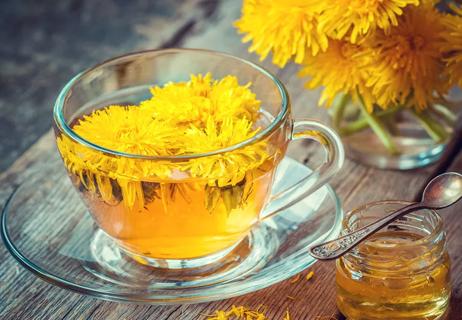 Dandelions steeping in hot water inside of a tea cup with a jar of honey in the foreground.