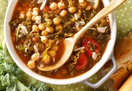 A bowl of chickpea soup made with kale and tomatoes with a wooden spoon in the bowl.