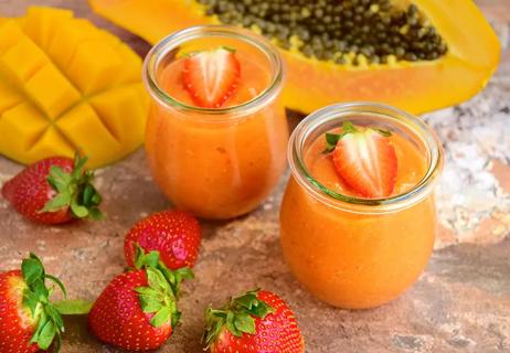 Two small glasses of mango strawberry smoothies with strawberry garnish displayed on a granite table with whole fruits