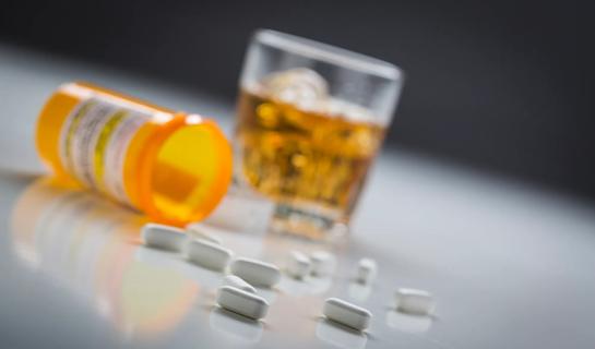 Open medicine bottle on its side, with spilled pills on the table, and glass of alcohol nearby