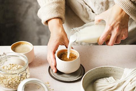 Person pouring homemade oat milk into mug of coffee, with squeezed oats and fresh oats in a jar nearby