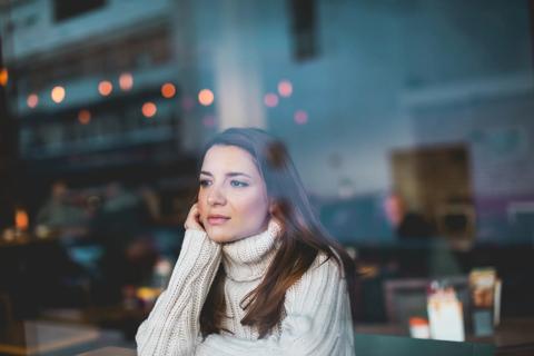 Person staring out cafe window looking sad, with reflecting holiday lights on window from busy street