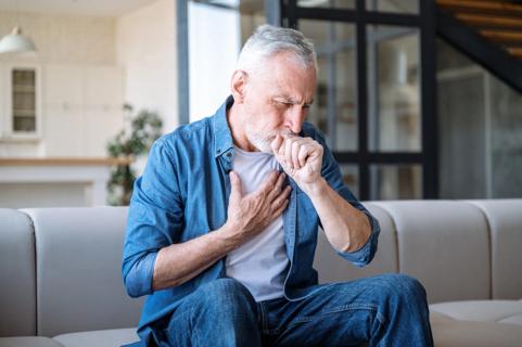 Older person sitting on couch, coughing, holding hand to chest