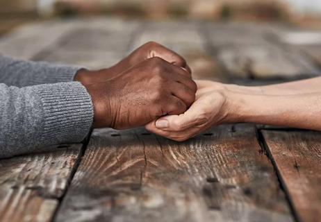 Two people hold hands in a comforting way across a wooden table.