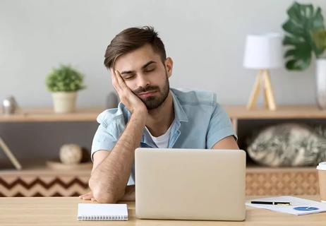 A person falls asleep while sitting at their laptop, leaning their head in their hand.