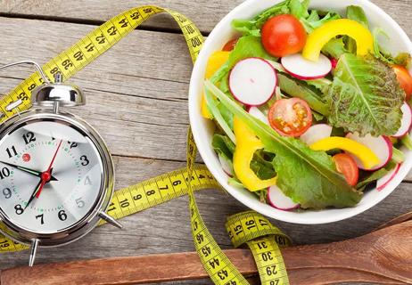 A salad, clock and measuring tape on a wooden tabletop.