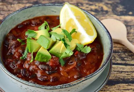 Closeup of meatless black bean chili in a stoneware bowl, with avocado and lemon garnish.