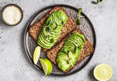 Two pieces of toast are on a plate, topped with sliced avocado, fennel seeds and lime.