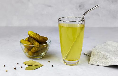 Glass of pickle juice with metal straw and glass bowl of pickles