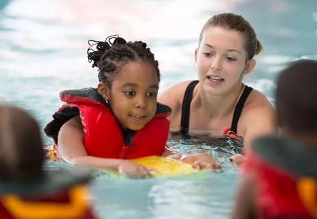 A child wearing a life jacket balances on a paddle-board with the help of a swimming instructor.