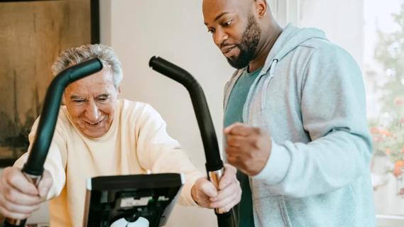 Man on an exercise bike with another man assisting