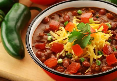 Closeup of Vegetarian Red Bean Chili in a red bowl placed on a wooden table with a pepper in the background.