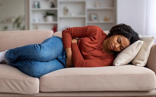 Person lying on couch, arms wrapped tightly around their stomach, grimacing