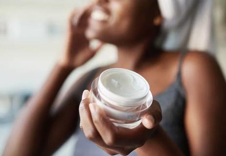 Closeup of a small pot of skin moisturizer and in background person applying it to their face.