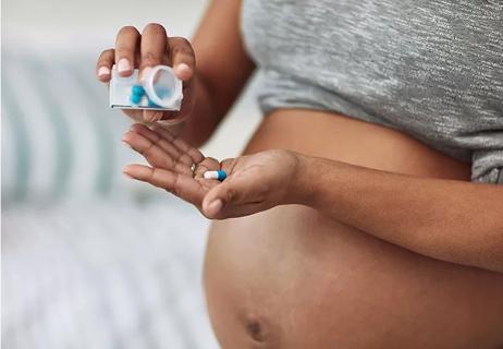 A pregnant person pours a blue and white pill into hand with shirt pulled up over bare belly