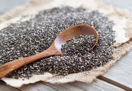 A wooden spoon scooping some chia seeds out of a mound of chia seeds in a burlap wrapper on a wooden table.